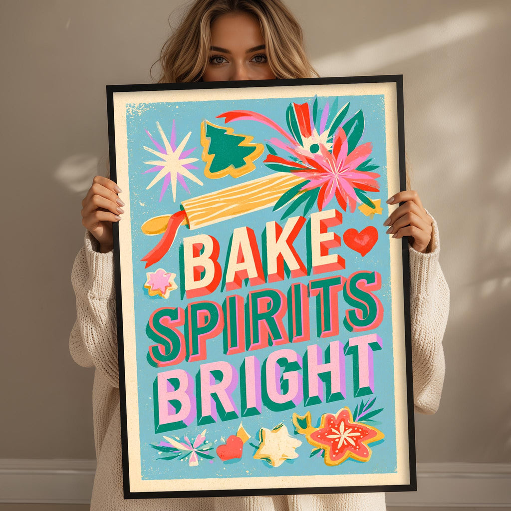 A woman is holding up a framed poster with the text "BAKE SPIRITS BRIGHT" and various colorful illustrations, including flowers, stars, and a starburst.