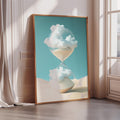 a large framed artwork depicting a glass hourglass with clouds inside, placed on a wooden floor against a white wall.