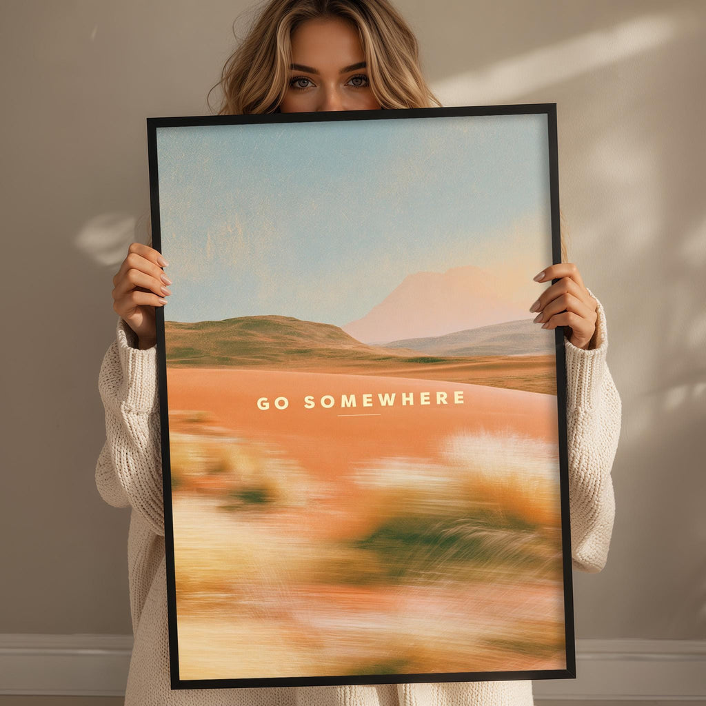 A woman holding a framed poster with a landscape scene and the text "GO SOMEWHERE" on it.