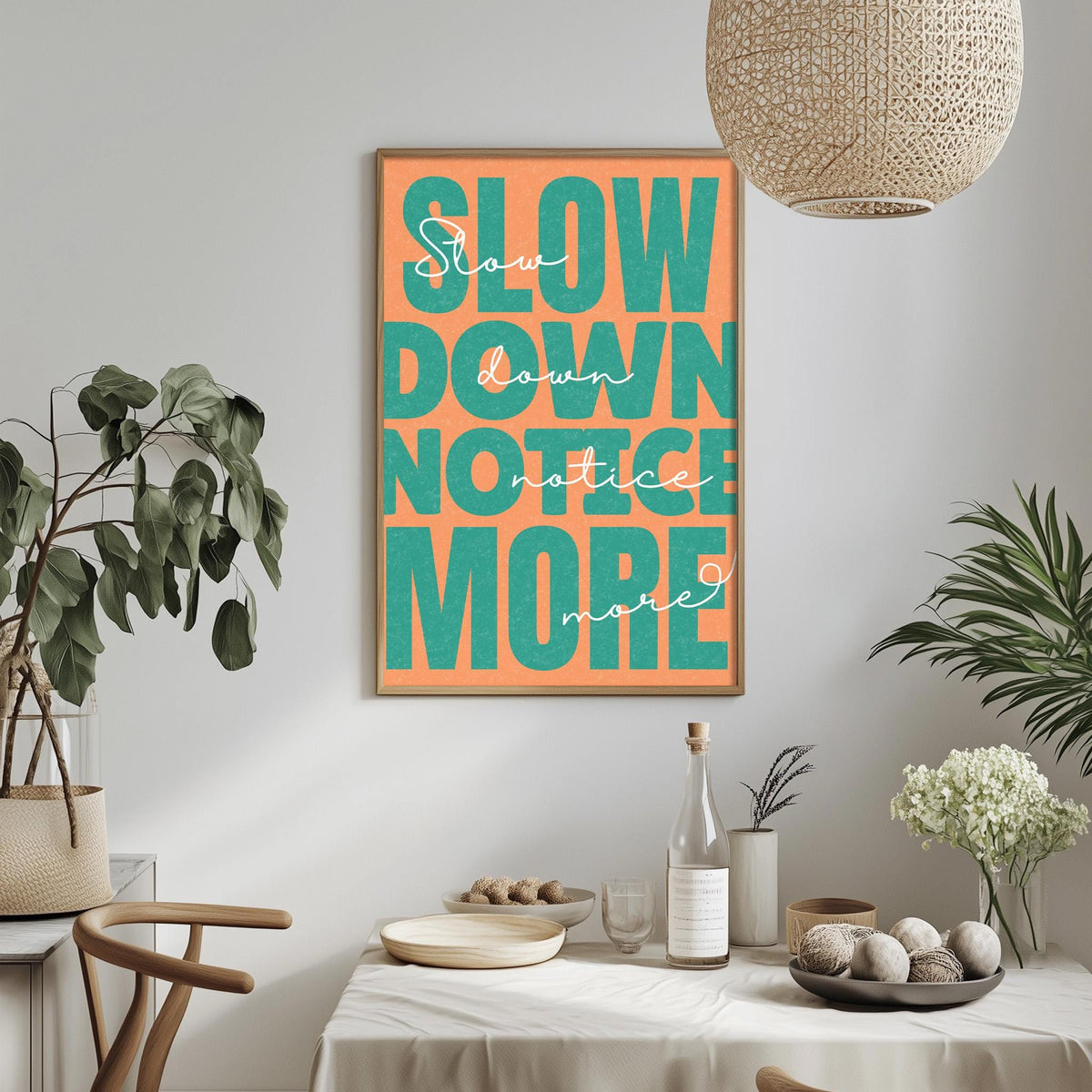 a dining table with a bowl of fruit, a bottle, and a vase, along with a potted plant and a hanging light fixture. Above the table, there is a framed poster with the text "Slow down, not more" in a vibrant orange and green color scheme.