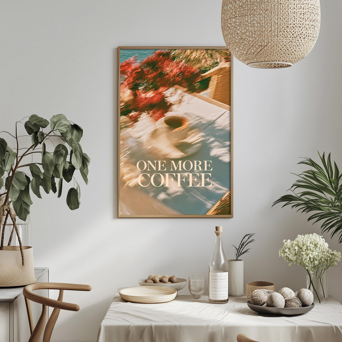 a dining table with a bowl of fruit, a bottle, and a vase, along with a potted plant and a hanging light fixture. Above the table, there is a framed poster with the text "ONE MORE COFFEE" displayed on it.
