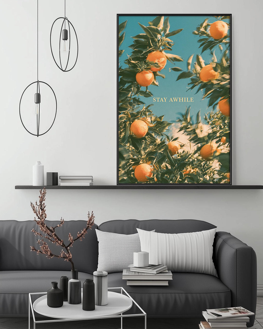 a cozy living room with a gray sofa, a coffee table, and a large framed poster of an orange tree on the wall. The poster has the text "STAY AWHILE" written on it.