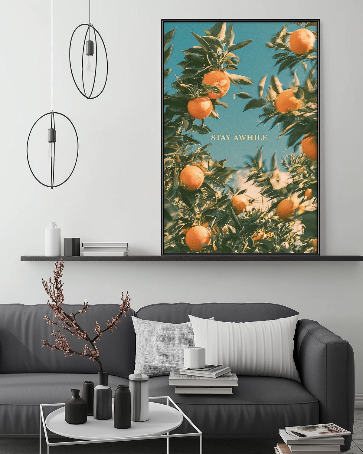 a cozy living room with a gray sofa, a coffee table, and a large framed poster of an orange tree on the wall. The poster has the text "STAY AWHILE" written on it.