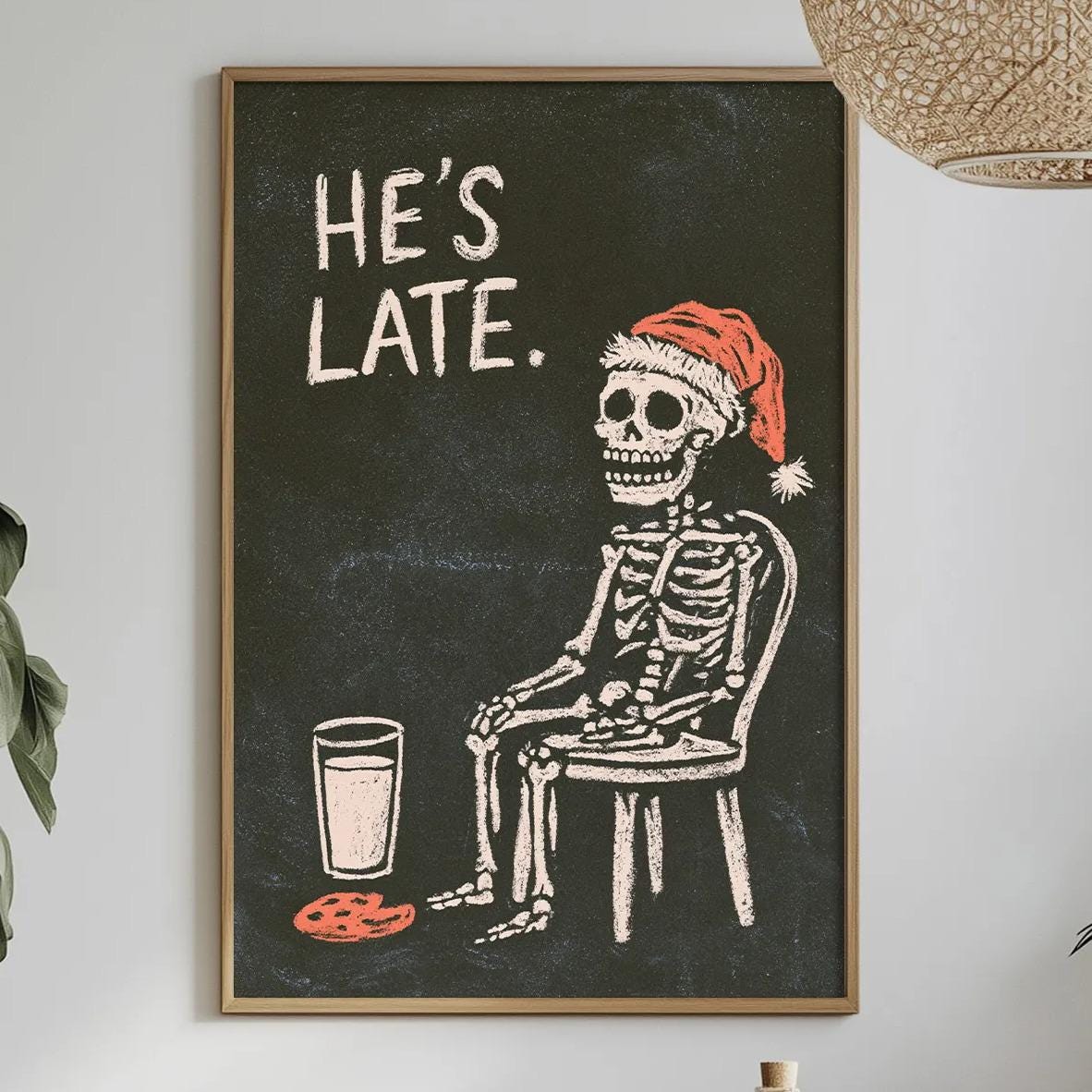 a skeleton wearing a Santa hat sitting in a chair, with a plate of cookies in front of him, and a framed poster of a skeleton with the text "HE'S LATE."