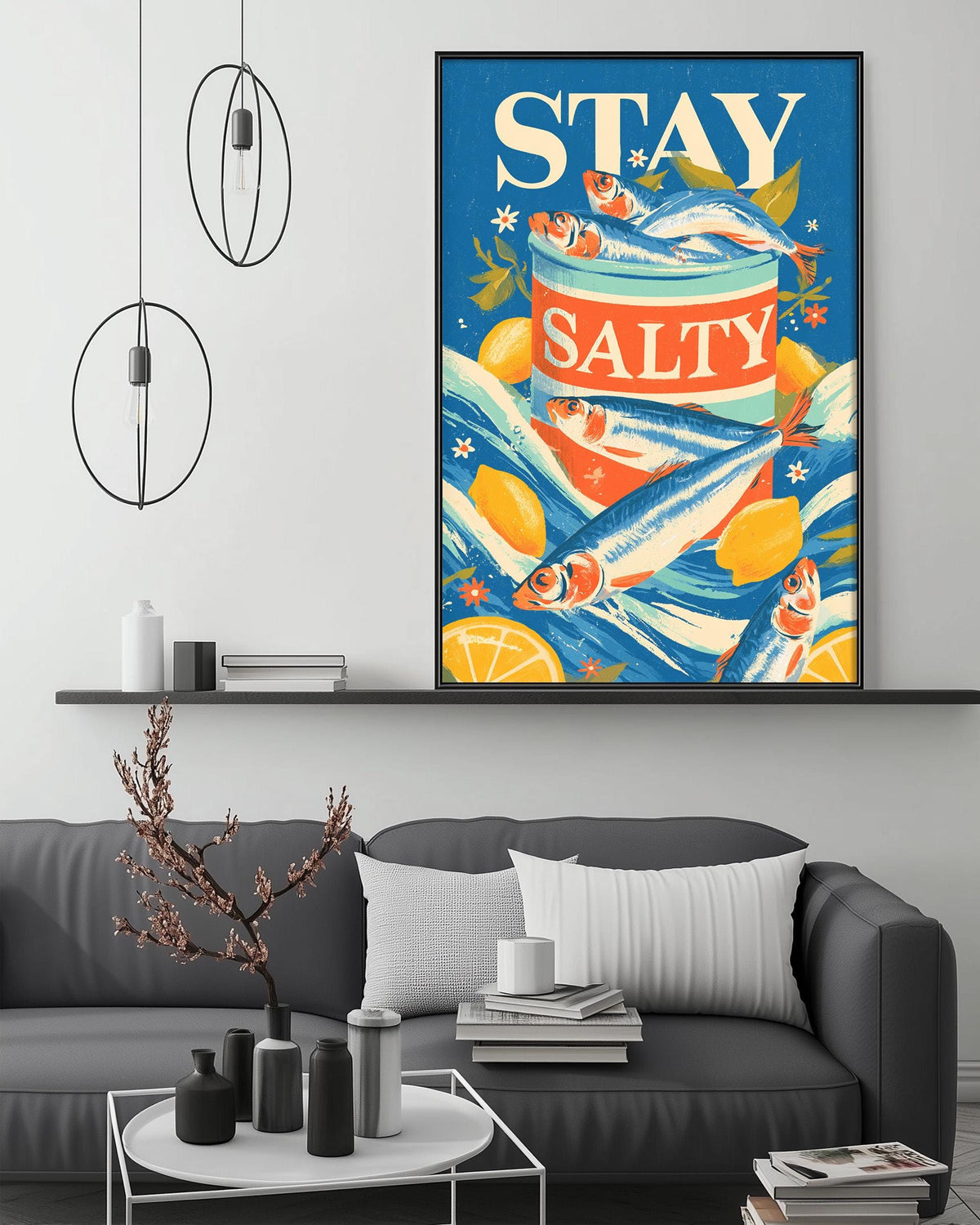 a large framed poster with the text "STAY SALTY" displayed prominently. The poster is set against a blue background with illustrations of fish and citrus fruits. The poster is hanging on a wall above a gray couch, which is positioned in front of a coffee table. The room has a minimalist and modern design, with a few decorative items such as vases and a potted plant.