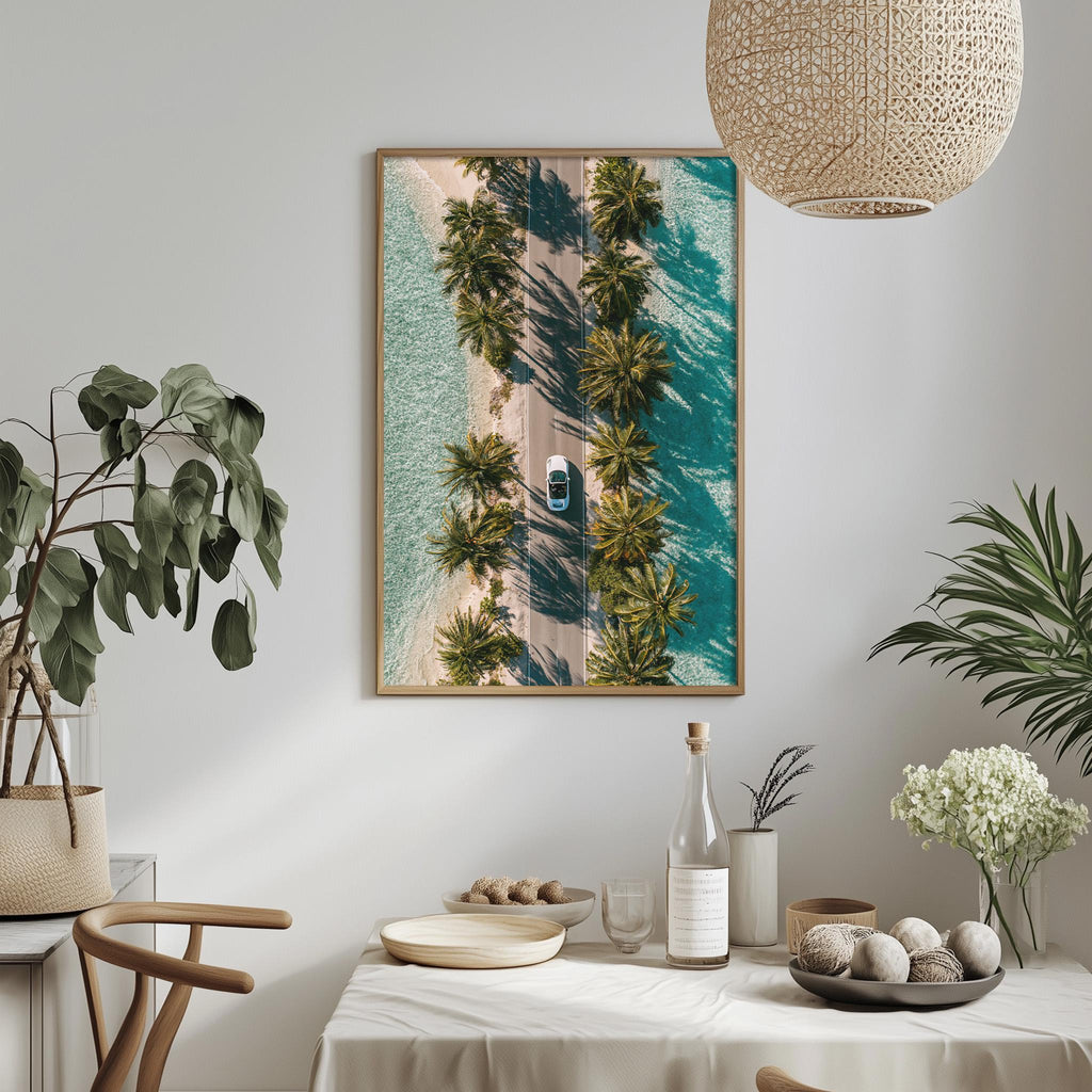 a dining table with a white tablecloth, a bottle, a bowl, and a vase with flowers. Above the table, there is a framed picture of a road lined with palm trees, and a hanging light fixture.