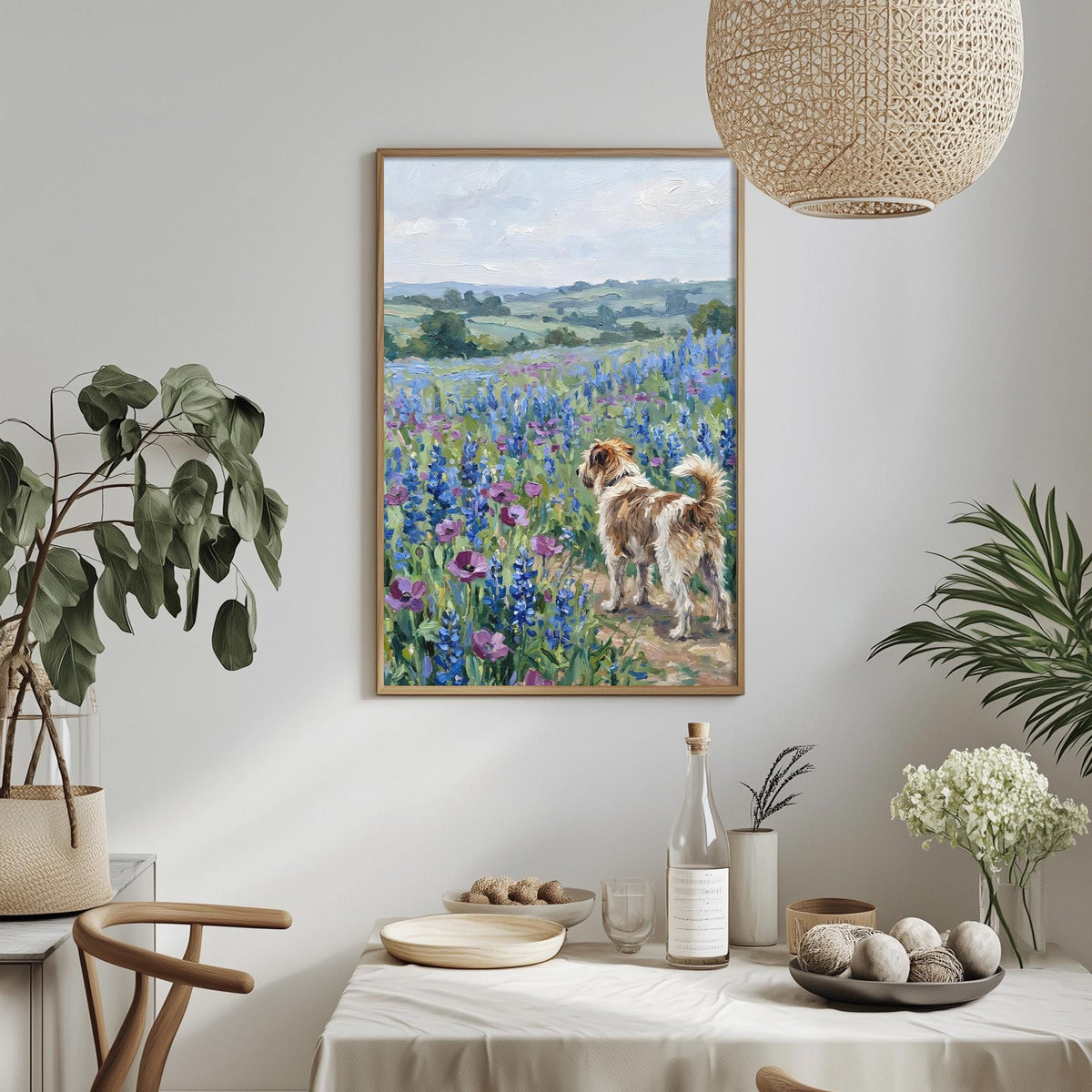 a painting of a dog standing in a field of flowers, with a serene landscape in the background. The painting is hung on a wall above a dining table, which has various items on it, including a bottle, a bowl, and a vase. The table is set with a white tablecloth and there are some plants and flowers in the room, creating a cozy and inviting atmosphere.
