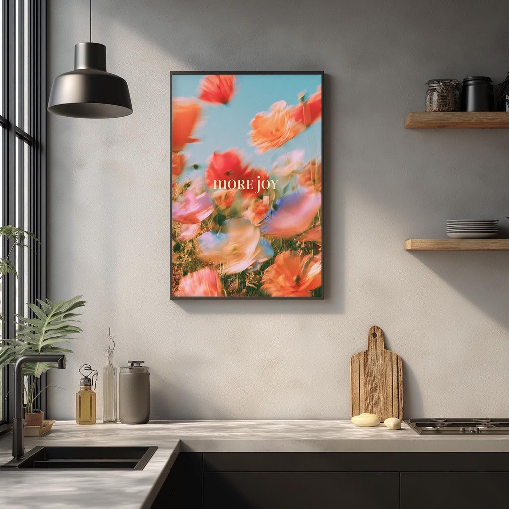 a kitchen counter with a framed poster of a field of flowers hanging above it. The poster has a vibrant, colorful image of flowers and the text "more joy" written on it. The kitchen counter also has various items such as a cutting board, a soap dispenser, and a bottle.