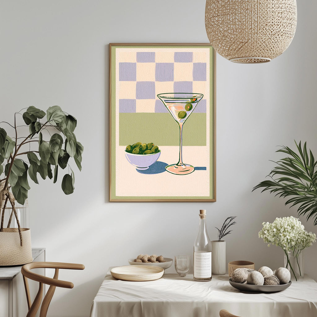 a dining table with a white tablecloth, a bowl of fruit, a bottle of wine, and a framed illustration of a martini glass with olives.