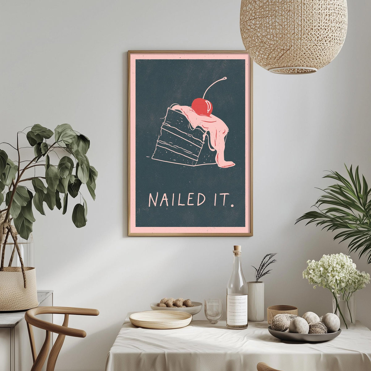 a framed poster with a whimsical illustration of a cake topped with a cherry, accompanied by the text "Nailed it." The poster is displayed on a wall above a dining table with various decorative items, including potted plants, vases, and a bowl of fruit.