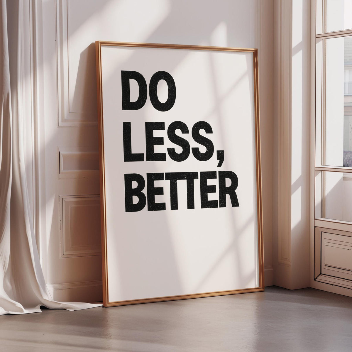 a large framed poster with the text "DO LESS, BETTER" on it, placed on a wooden floor in a room with a window.