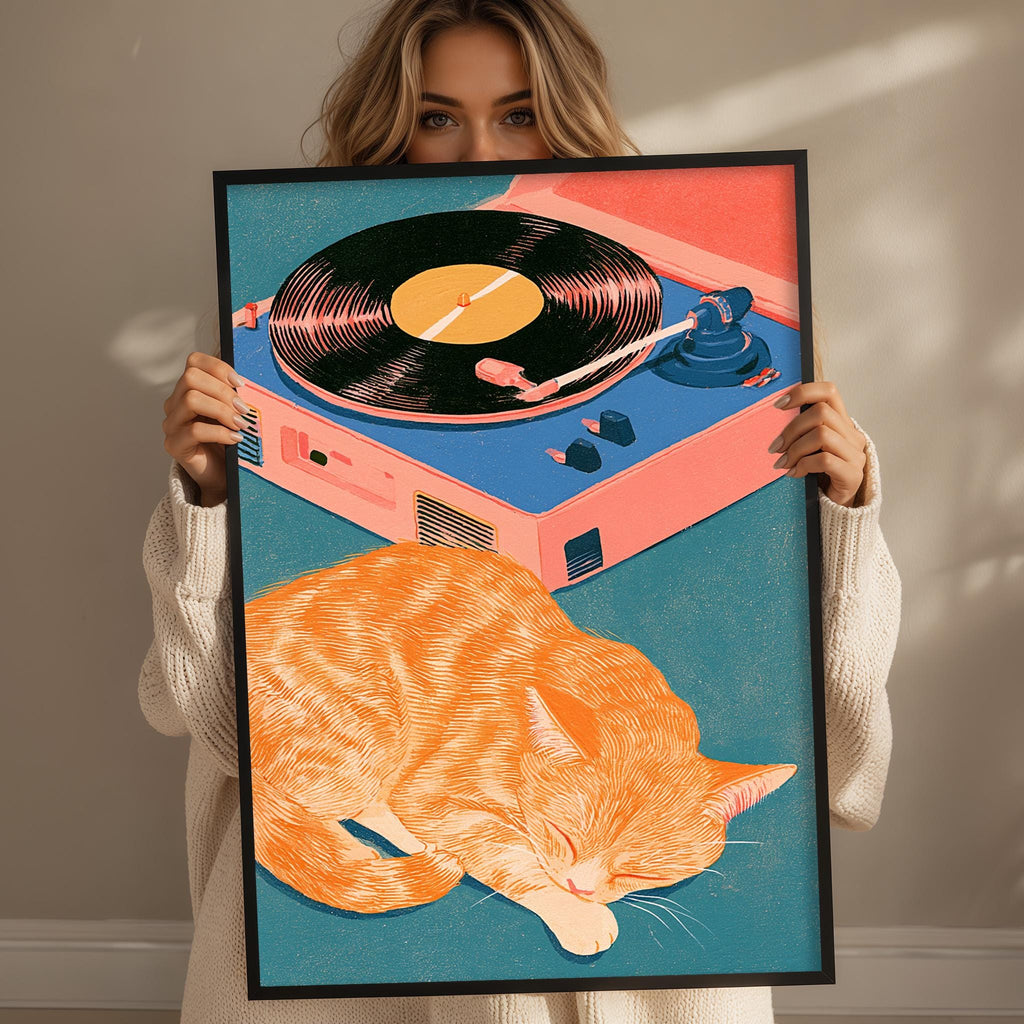 A woman holds up a framed illustration of a sleeping orange cat next to a record player.