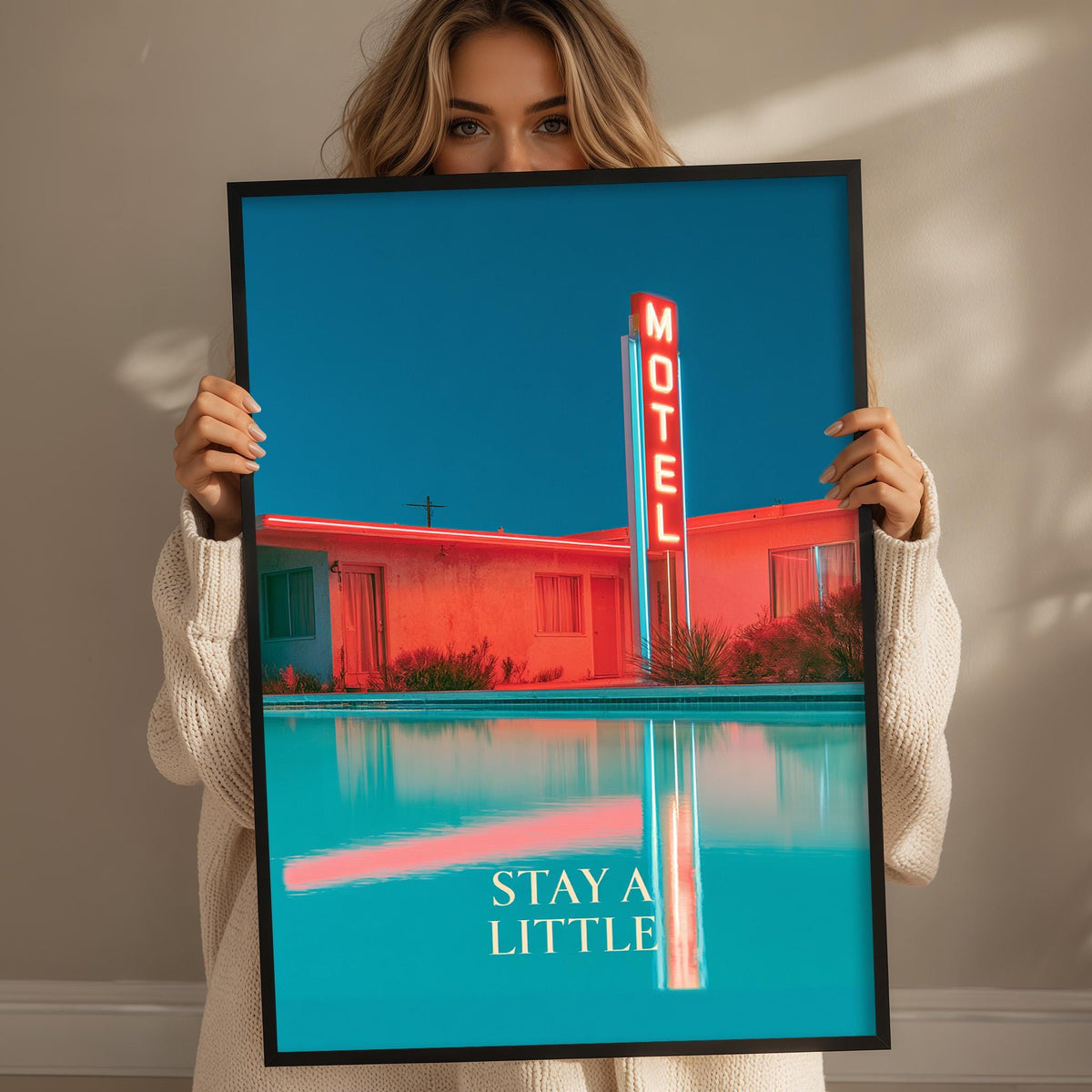 A woman holds up a framed poster with a vibrant motel sign and a pool, with the text "STAY A LITTLE" displayed on the poster.