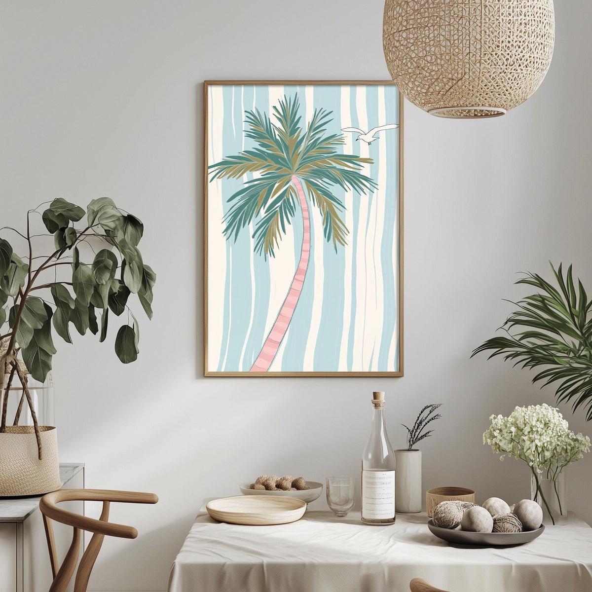 a dining table with a white tablecloth, a bowl of fruit, a bottle, and a vase with flowers. Above the table, there is a framed picture of a palm tree with a bird in flight.