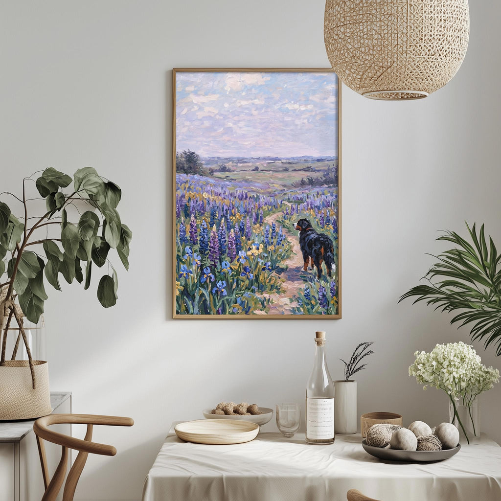 a dining table with a white tablecloth, a vase of white flowers, a bottle of wine, and a bowl of fruit. Above the table, there is a framed painting depicting a field of purple flowers with a dog walking through it.