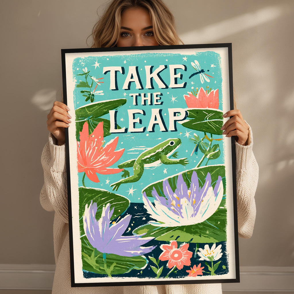 A woman is holding up a framed poster with a vibrant illustration of a frog, flowers, and lily pads.