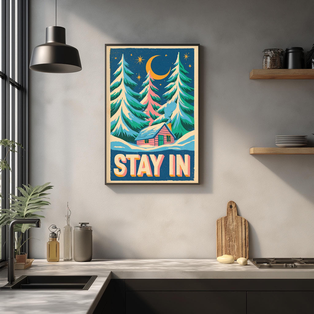 a kitchen counter with a framed poster hanging on the wall above it. The poster depicts a snowy landscape with pine trees and a cabin, along with the text "STAY IN" prominently displayed.