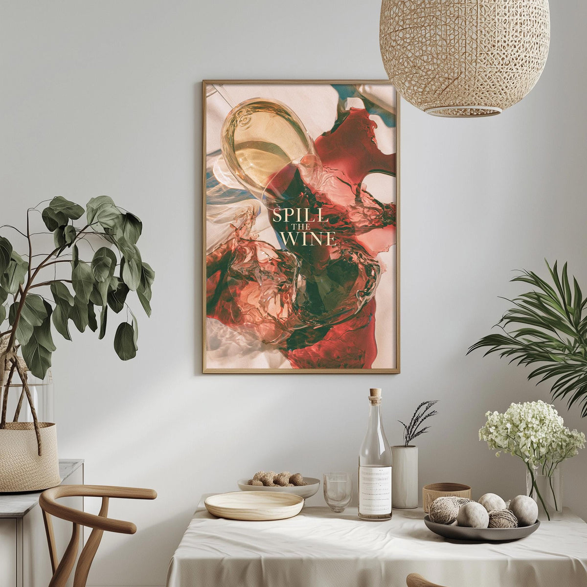 a dining table with a white tablecloth, a bowl of fruit, a bottle of wine, and a plant. Above the table, there is a framed poster with the text "SPILL THE WINE" in bold letters.