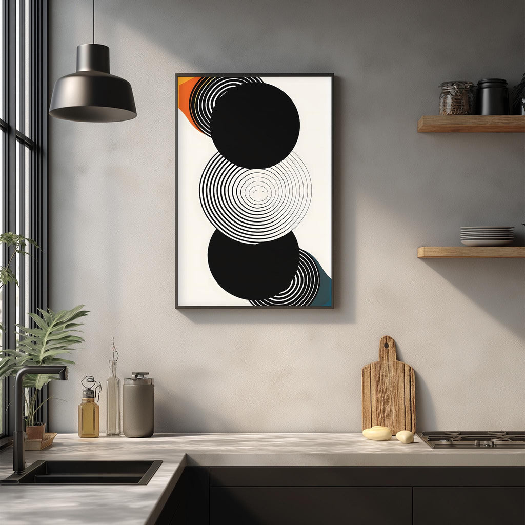a modern kitchen with a minimalist design. The focal point is a framed abstract artwork with black and white circles and a wooden cutting board on the countertop. The kitchen has a sleek, clean aesthetic with a black countertop, white walls, and a pendant light above the sink.