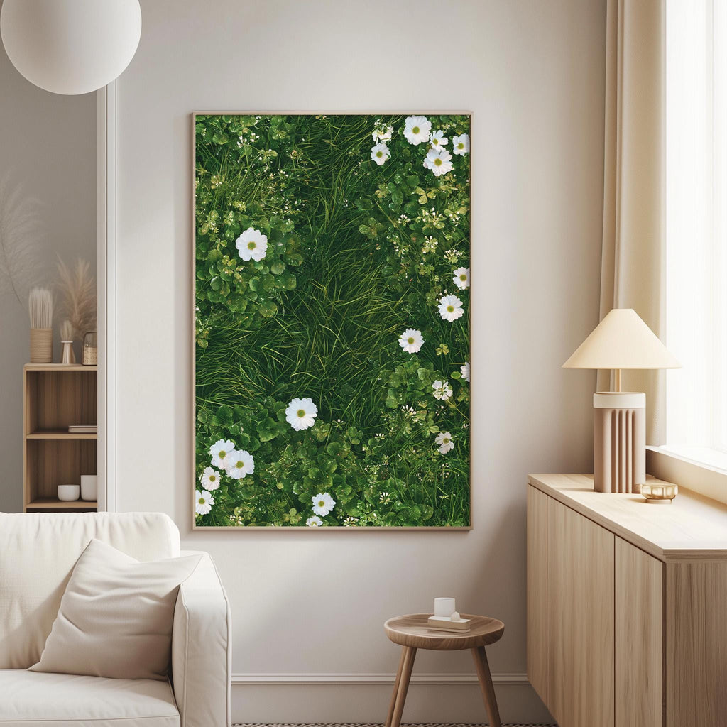 a serene living room with a large framed picture of a lush green grassy area filled with white flowers hanging on the wall. The room is furnished with a white sofa and a wooden side table, creating a cozy and inviting atmosphere.