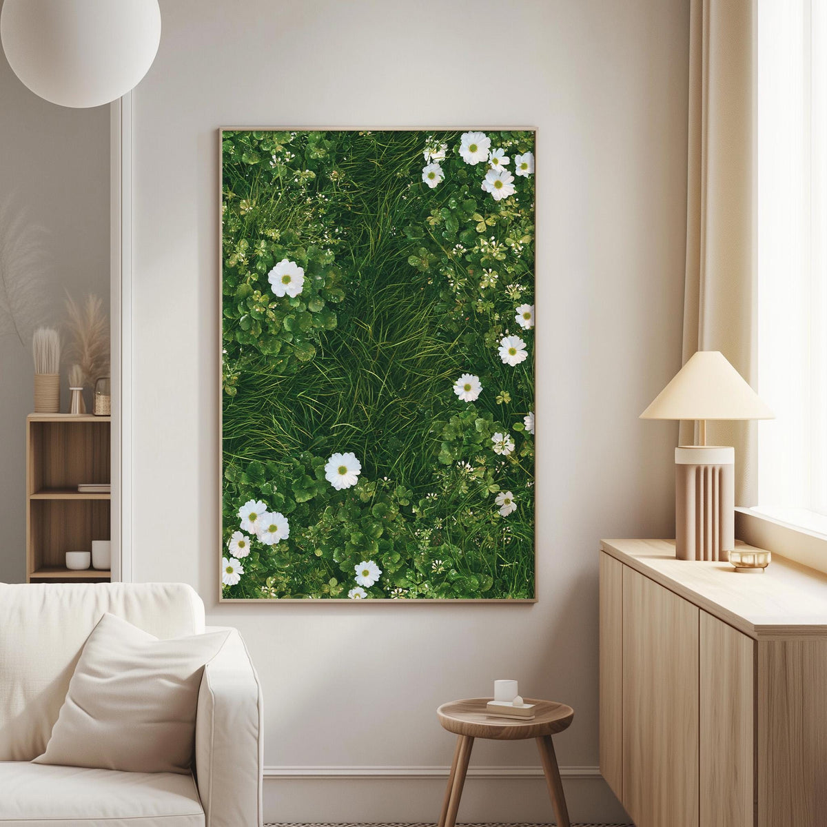 a serene living room with a large framed picture of a lush green grassy area filled with white flowers hanging on the wall. The room is furnished with a white sofa and a wooden side table, creating a cozy and inviting atmosphere.