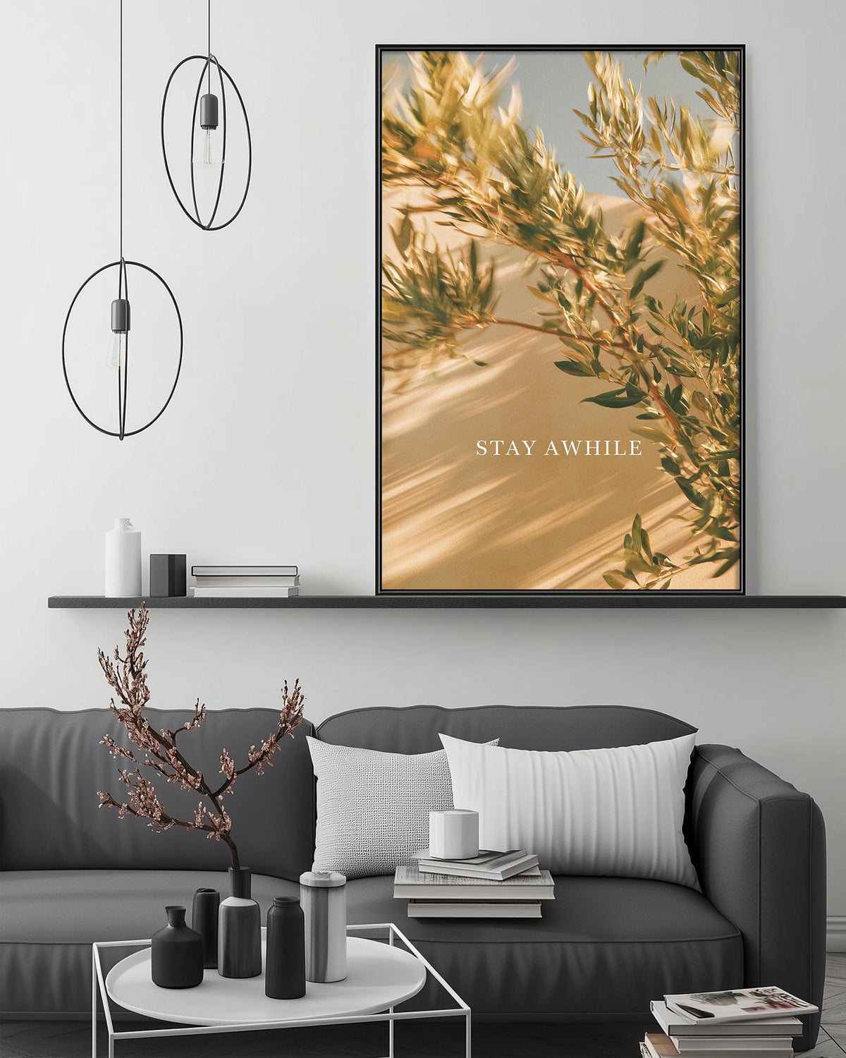 a modern living room with a gray sofa, a coffee table, and a large framed print of a desert landscape with a tree branch. The print has the text "STAY AWHILE" written on it.