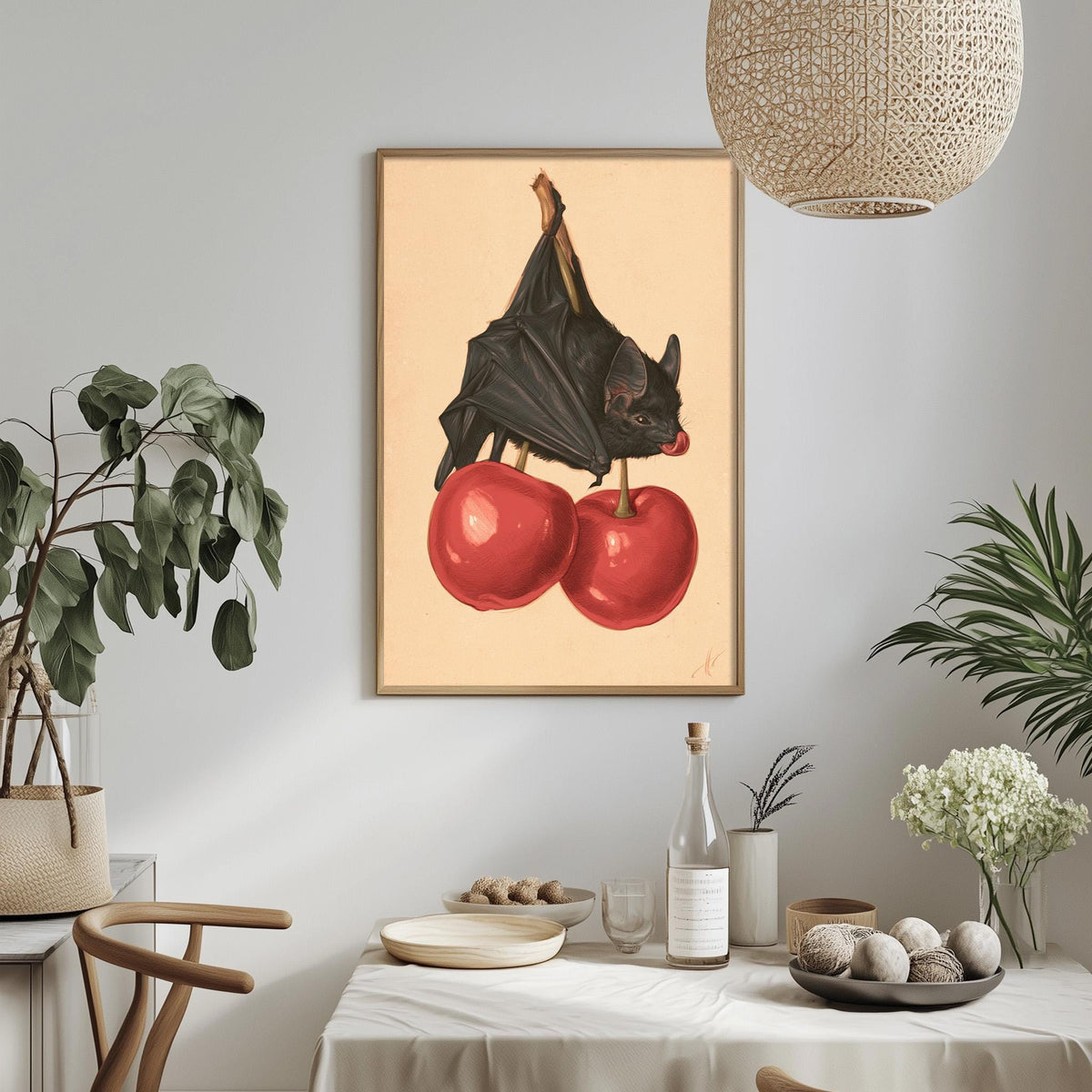 a framed artwork of a bat perched on two red cherries, hanging on a wall above a table with various items on it.