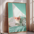 A framed image of a cat wearing pink sunglasses, lying on a bed next to a clock.