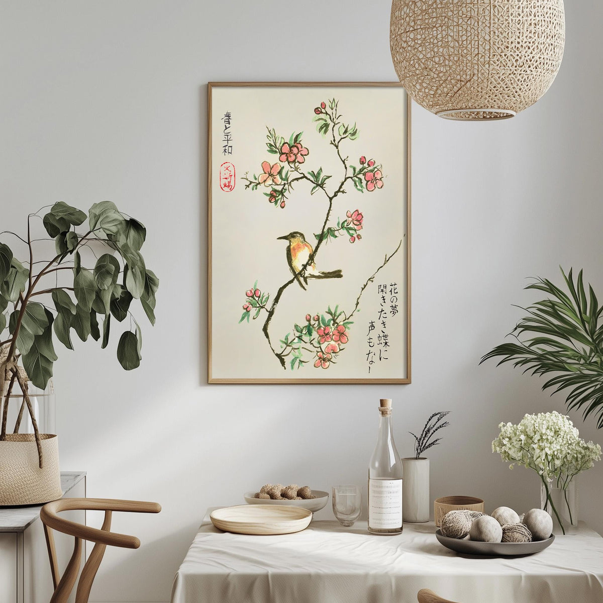 a dining table with a white tablecloth, a bowl of fruit, a bottle, and a vase with flowers. Above the table, there is a framed picture of a bird on a branch with flowers. The table is set with a plate, a cup, and a bowl. The room has a minimalist and elegant design, with a hanging light fixture and potted plants adding to the decor.