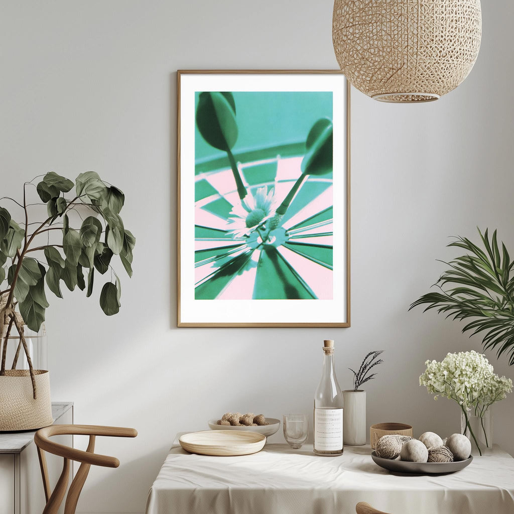 a dining table with a white tablecloth, a bowl of fruit, a bottle, and a vase with flowers. Above the table, there is a framed picture of a dartboard with a green and pink color scheme.
