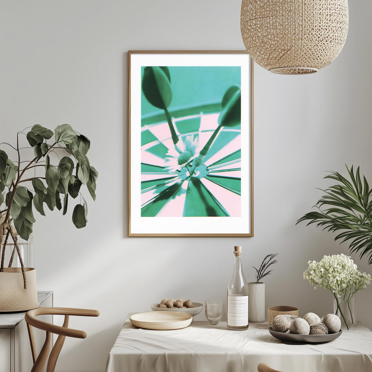 a dining table with a white tablecloth, a bowl of fruit, a bottle, and a vase with flowers. Above the table, there is a framed picture of a dartboard with a green and pink color scheme.