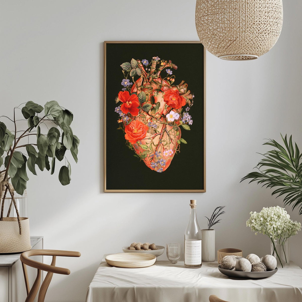 a dining table with a white tablecloth, a vase of white flowers, a bottle, and a bowl of fruit. Above the table, there is a framed picture of a floral heart on the wall.