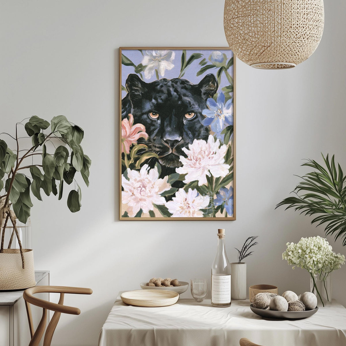 a dining table with a white tablecloth, a vase of white flowers, a bottle, and a bowl of fruit. Above the table, there is a framed painting of a black panther surrounded by pink and white flowers.