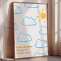 A framed poster with a drawing of clouds and a sun on a white background, placed on a wooden floor next to a window.