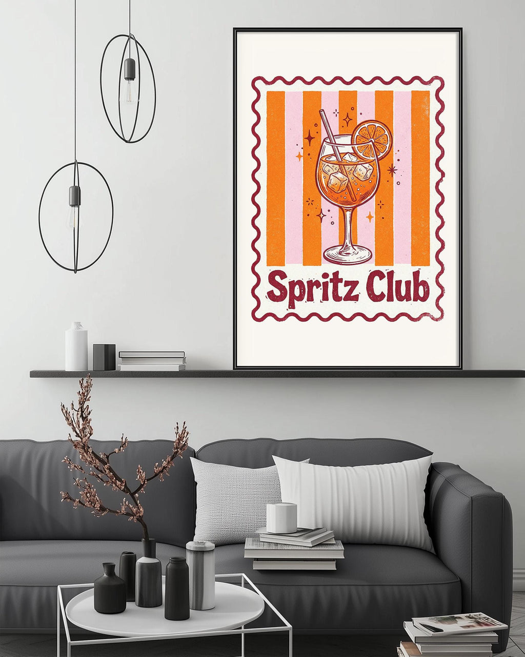 a framed poster of a glass of orange juice with a lemon slice, accompanied by the text "Spritz Club" in a decorative border. The poster is displayed on a wall above a gray couch, which has a coffee table in front of it. The room also contains a shelf with various decorative items, and a potted plant is visible in the background.