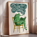 a framed illustration of a green frog sitting in a chair and reading a book, with a cloud-like shape above the frog.