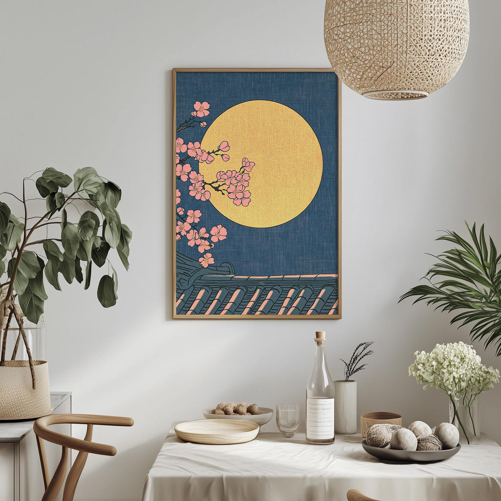 a dining table with a white tablecloth, a bowl of fruit, a bottle, and a vase with flowers. Above the table, there is a framed artwork depicting a night scene with a large yellow moon and pink flowers.