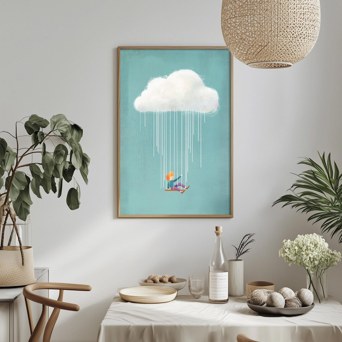 a dining table with a white tablecloth, a bowl of fruit, a bottle, and a vase with flowers. Above the table, there is a framed picture of a person sitting on a swing under a cloud, with rain falling from it.