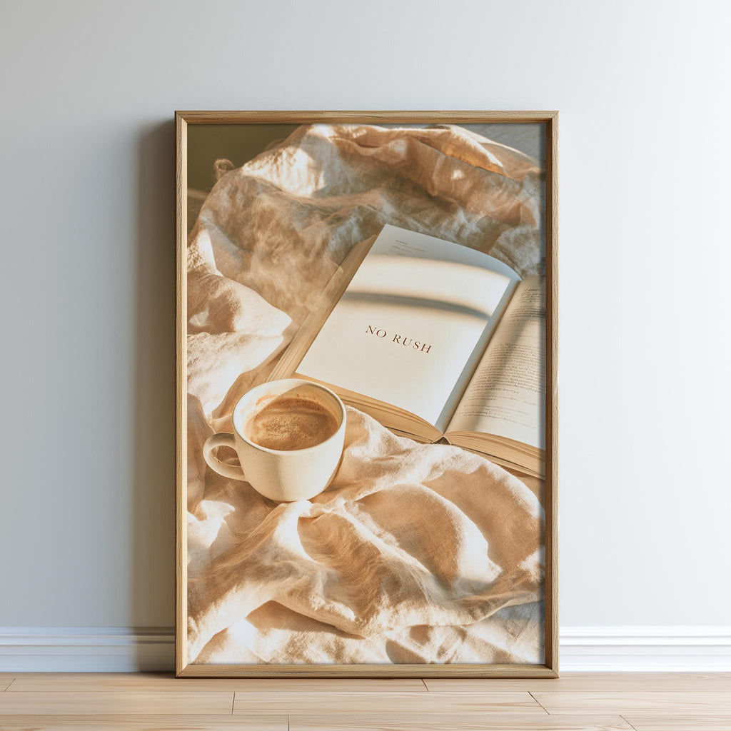 A framed image of an open book with the text "NO RUSH" on the cover, accompanied by a cup of coffee, placed on a beige blanket against a white wall.