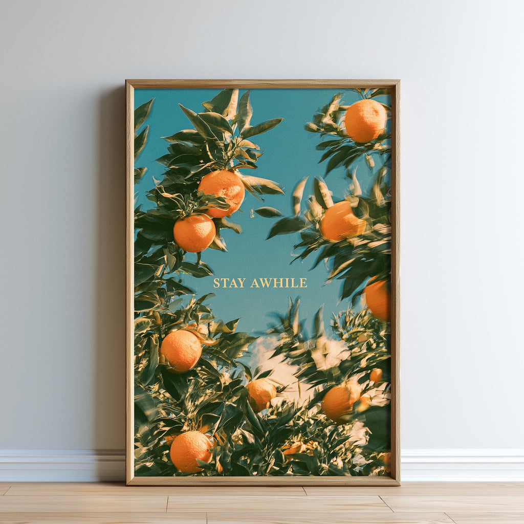 The image is a framed poster featuring a close-up view of an orange tree with ripe oranges hanging from its branches. The text "STAY AWHILE" is displayed in the center of the poster.