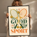 A woman holding a framed poster with the text "GOOD SPORT" and tennis rackets, flowers, and stars on it.