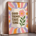 A framed poster with a colorful design and the text "GOOD THINGS TAKE TIME" on it, placed on a wooden floor next to a window.