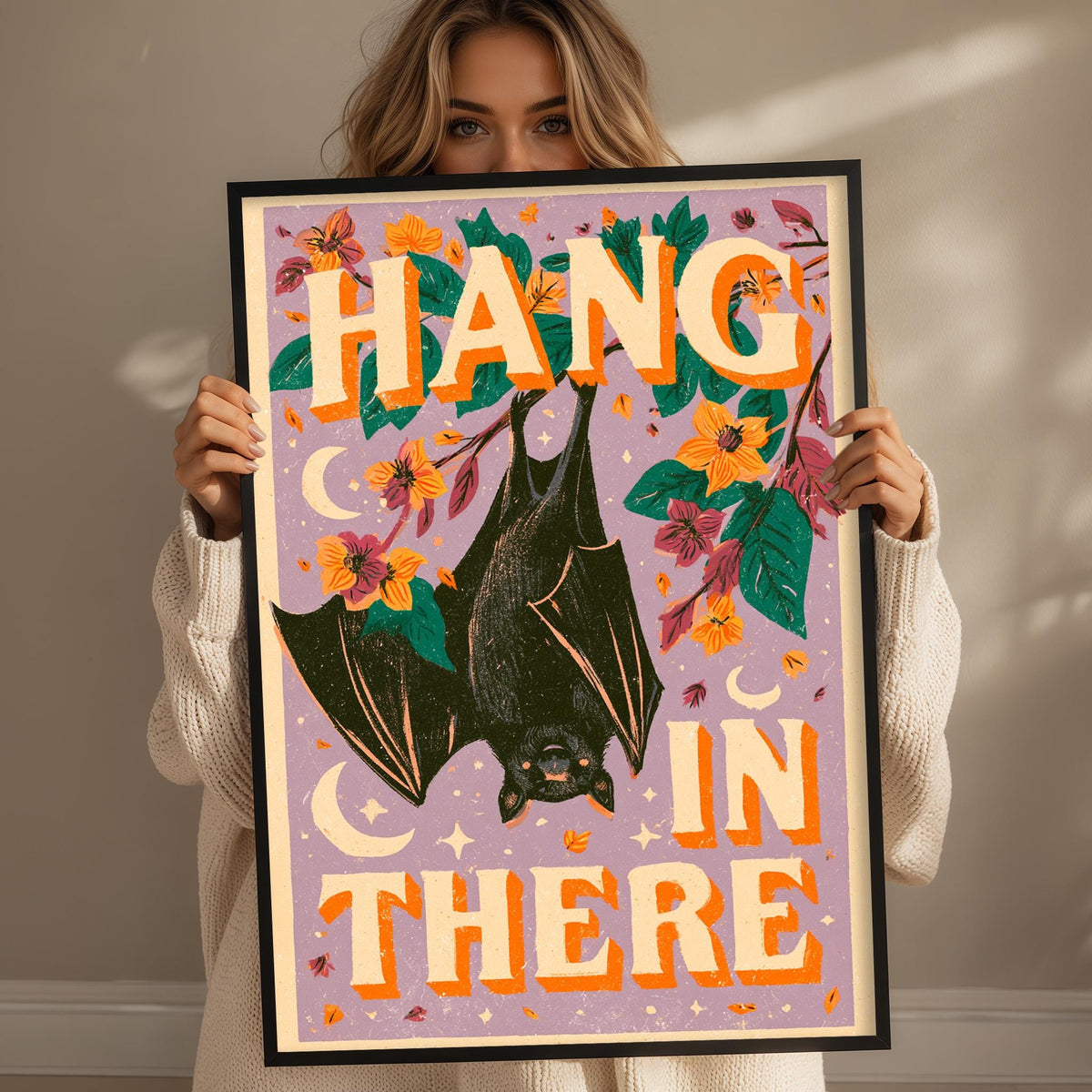 A woman is holding up a framed poster with a bat illustration and the text "HANG IN THERE" written on it.