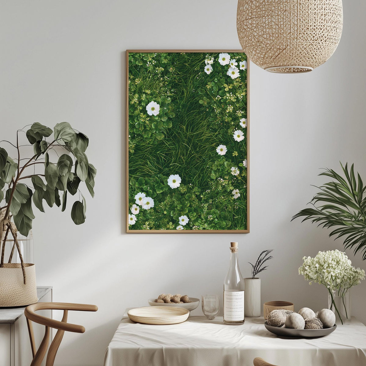 a dining table with a white tablecloth, a bowl of fruit, a bottle, and a vase with flowers. Above the table, there is a framed picture of a lush green field with white flowers. The table is set with a bottle, a wine glass, and a plate. The room has a minimalist and modern design, with a woven light fixture hanging above the table.