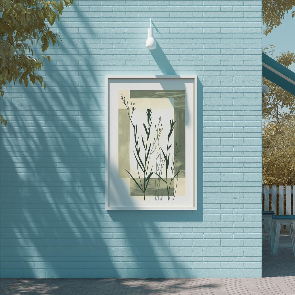 a blue brick wall with a framed artwork of green plants hanging on it. The artwork is framed in white and is displayed on the wall, creating a visually appealing contrast between the natural elements and the urban setting.