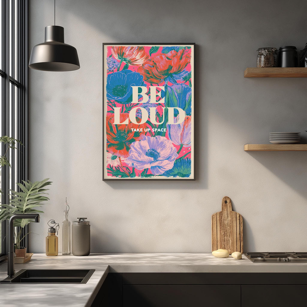 a kitchen counter with a framed poster hanging on the wall above it. The poster has a colorful floral design and the text "BE LOUD TAKE UP SPACE" written on it.