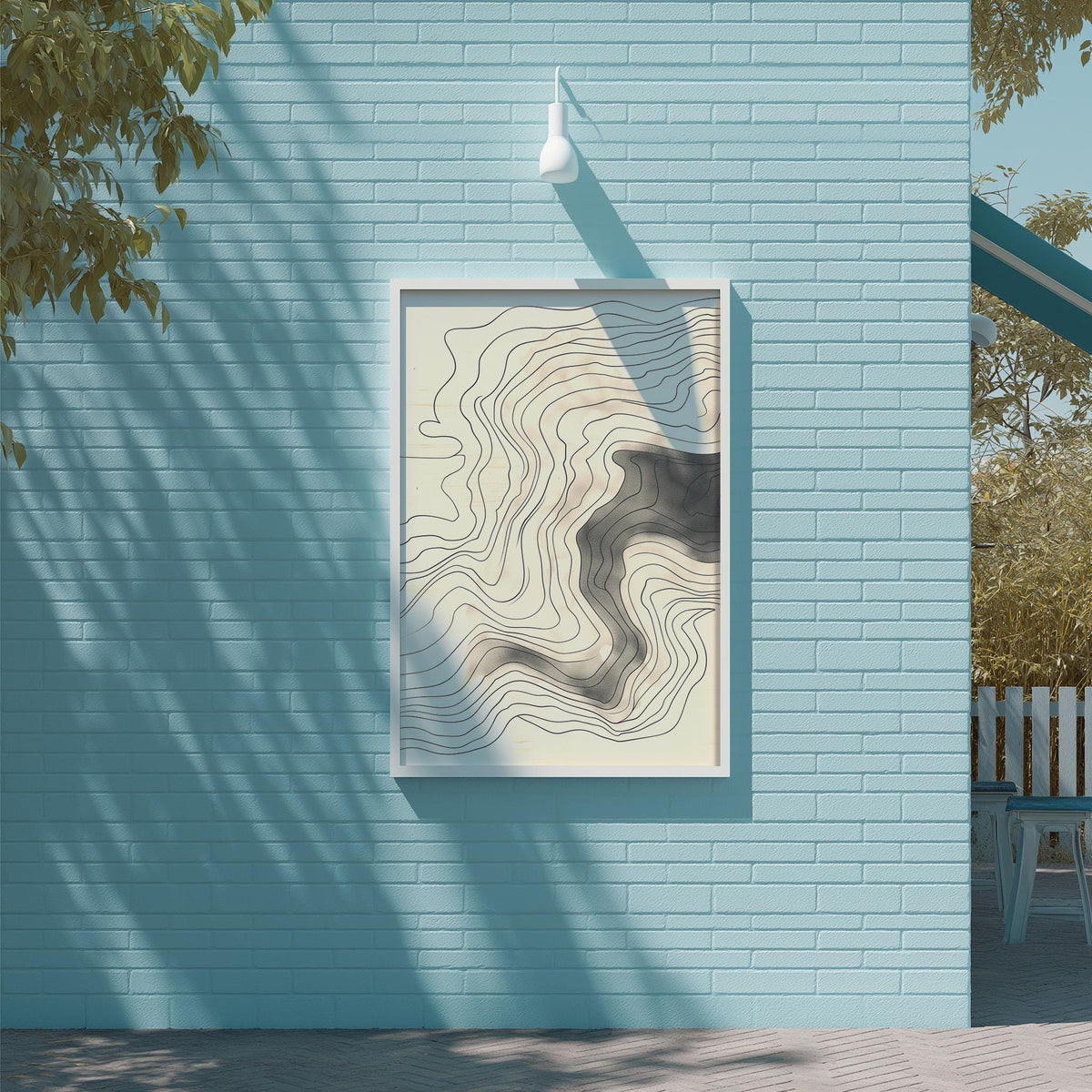 a blue brick wall with a framed abstract artwork hanging on it. The artwork has a wavy, abstract design in shades of gray and white. The wall is situated next to a tree, and there is a white light fixture mounted on the wall.