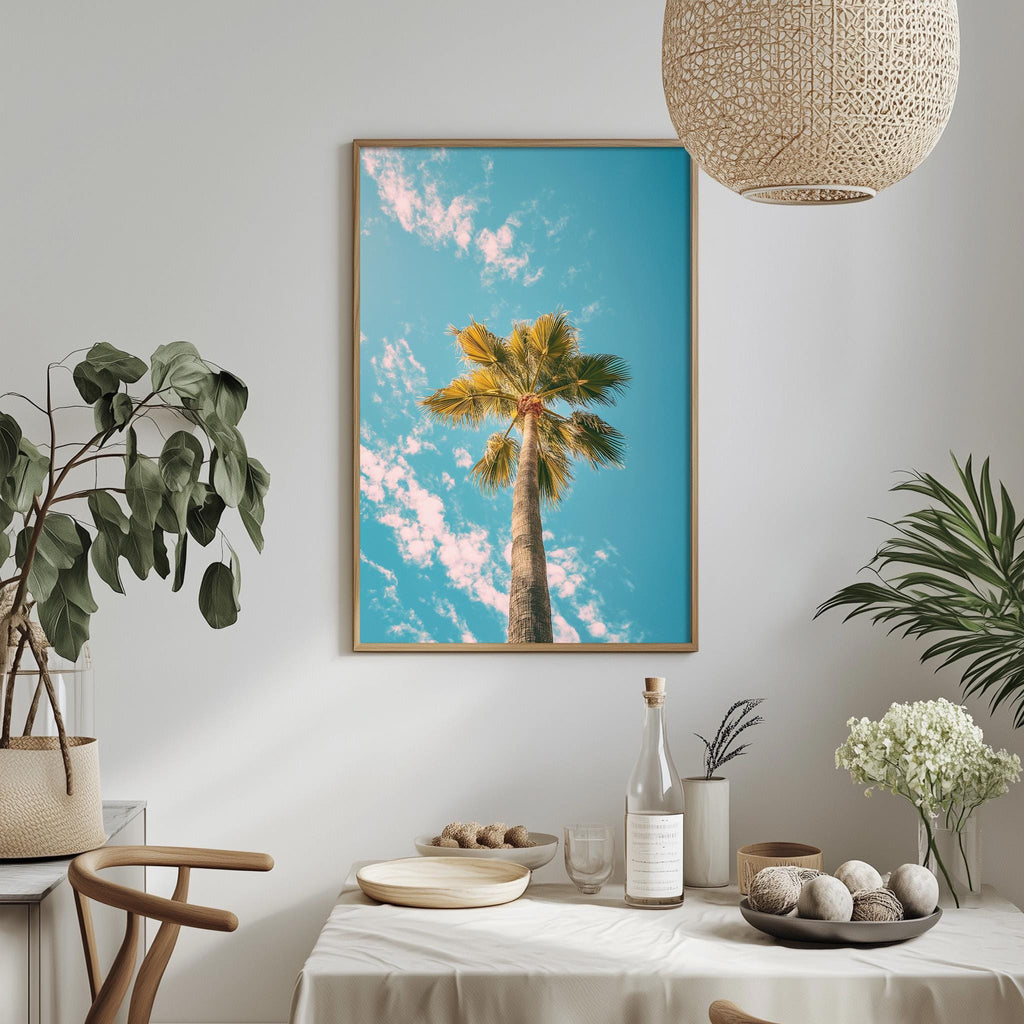a dining table with a white tablecloth, a bowl of fruit, a bottle, and a vase with flowers. Above the table, there is a framed picture of a palm tree against a blue sky with clouds.