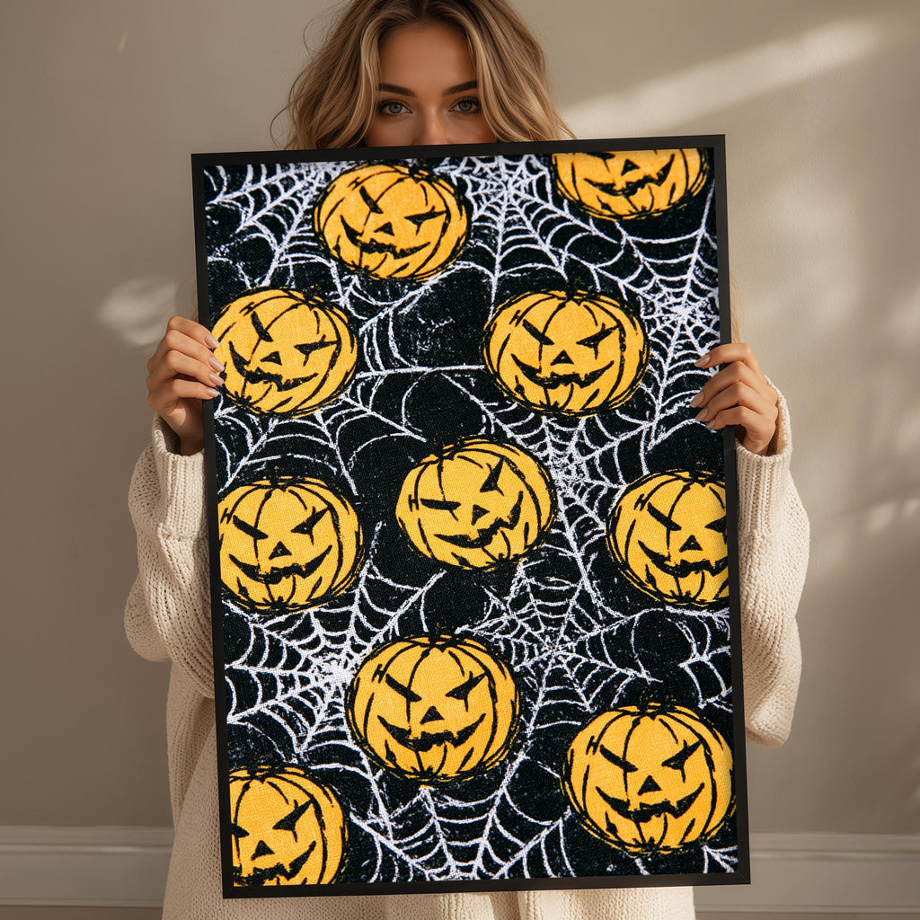 A woman holds up a framed print with a Halloween-themed design featuring multiple jack-o-lanterns and a spider web pattern.