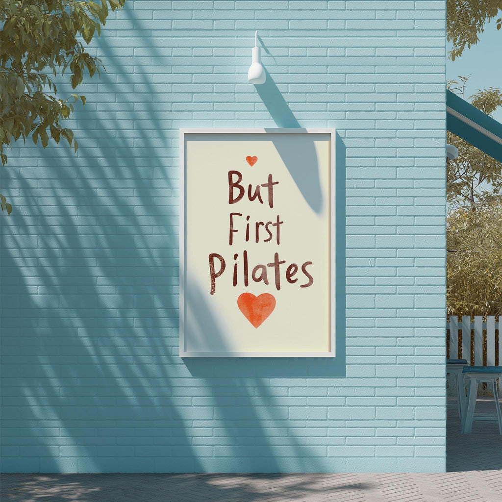 A framed poster with the text "But First Pilates" and an orange heart symbol is displayed on a blue brick wall.